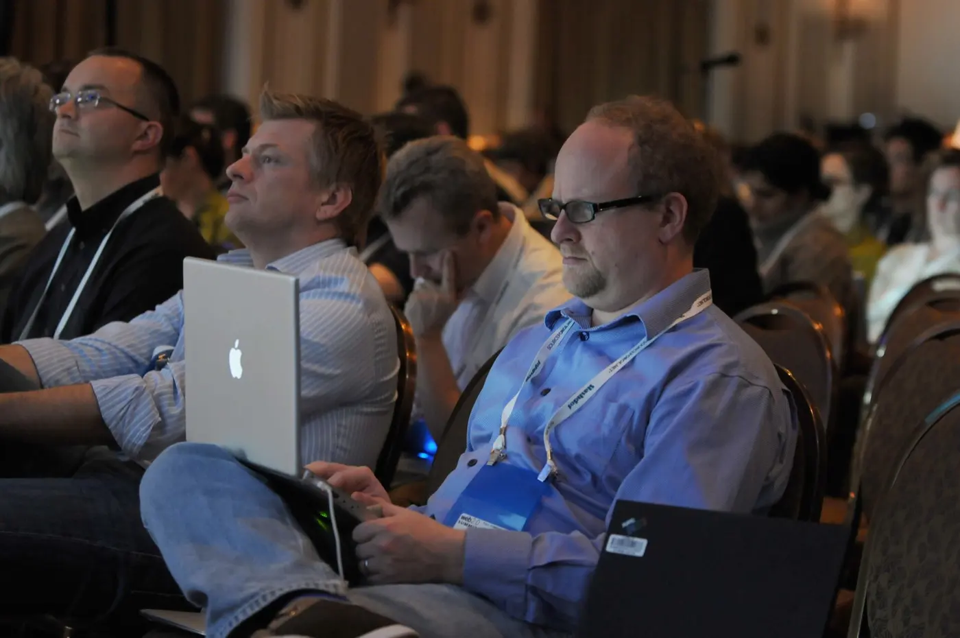 An audience at a conference, several with laptops open